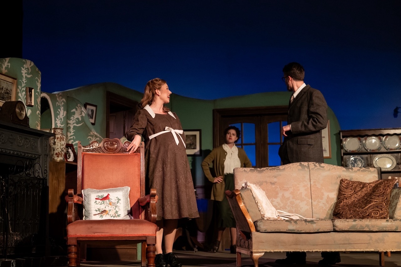 Theatre scene with woman in brown dress standing on chair, man in dark suit, and third person in background on stage with furniture.