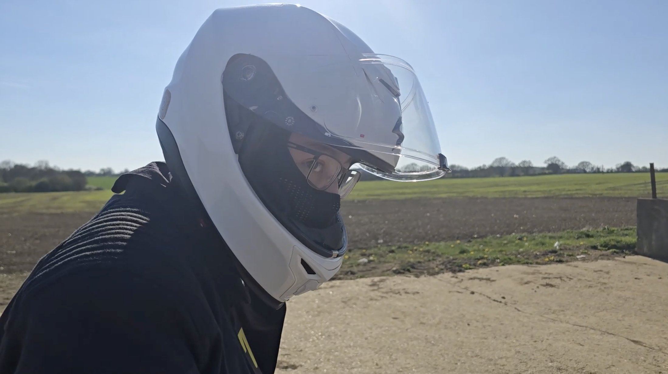 Person wearing white motorcycle helmet with visor, black jacket, standing on dirt path with green fields and blue sky behind.