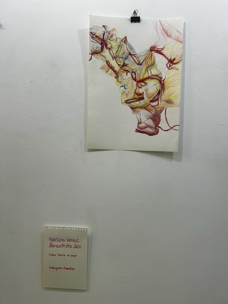 An abstract mixed-media artwork clipped to a white wall, with a small label mounted below it.