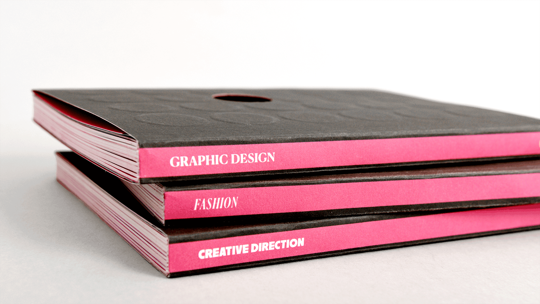 Stack of three books with bright pink spines and black covers. White text on spines reads "GRAPHIC DESIGN", "FASHION", and "CREATIVE DIRECTION".