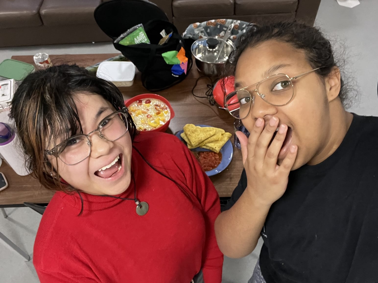 Two students taking a selfie, one with their hand over their mouth and the other smiling with a red sweater, food and brown sofa in the background.