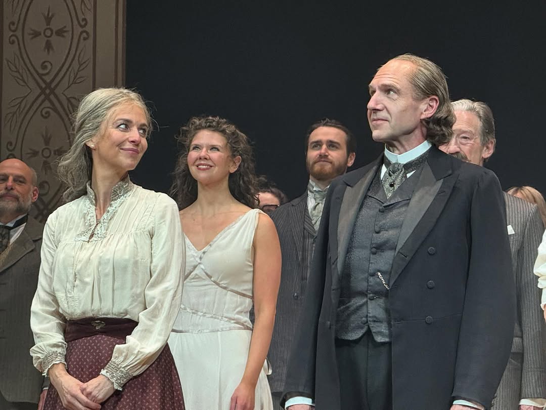 A group of actors on stage, including Miranda Raison on the left and Ralph Fiennes on the right. The woman stands to the left, wearing a white blouse and a brown skirt, while a young woman in a white dress is positioned centre. The backdrop is patterned, and several other actors are visible in the background.