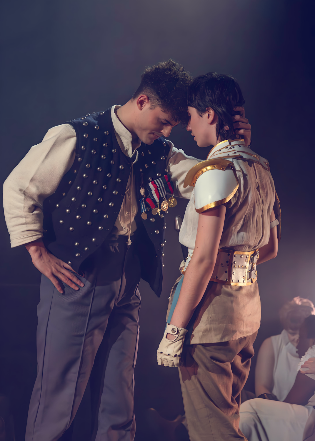 A young man in a decorated vest and dark trousers stands facing a young woman wearing light clothing and protective gear. They are in a dimly lit stage setting, with other figures partially visible in the background.