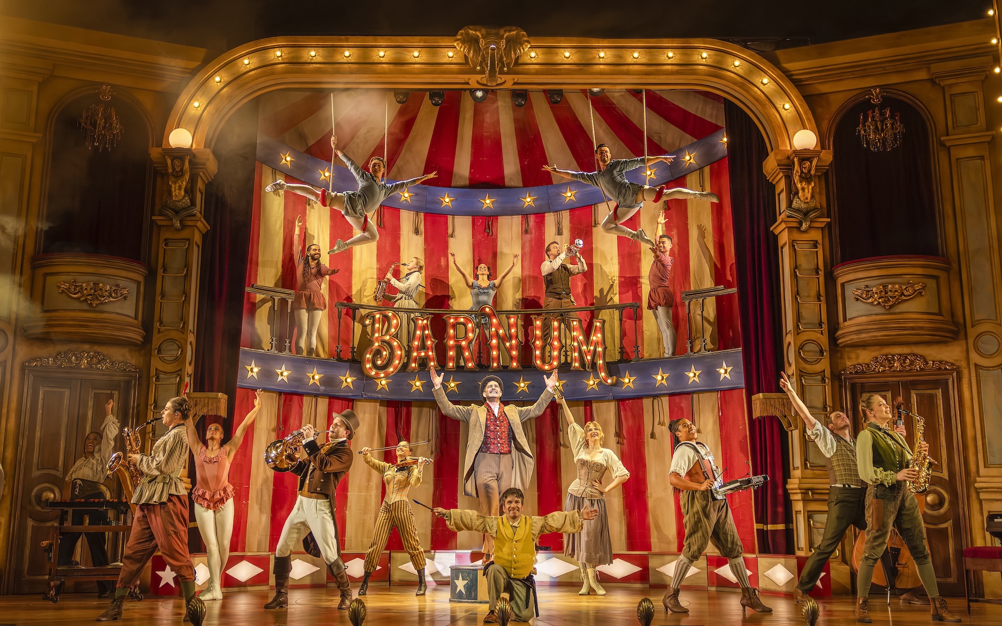 A vibrant theatre stage set for a performance of 'Barnum', featuring actors in colourful costumes. The backdrop displays red and white stripes with 'BARNUM' illuminated in lights. Performers are arranged in groups, showcasing various acts.