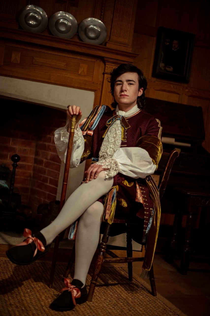 A stately man dressed in 17th-century clothing sits in a dark parlour. He is wearing a rich suit of wools and velvets, decorated with lace and festoons of ribbons. One leg is hooked over his knee and he holds onto a cane, looking proud.