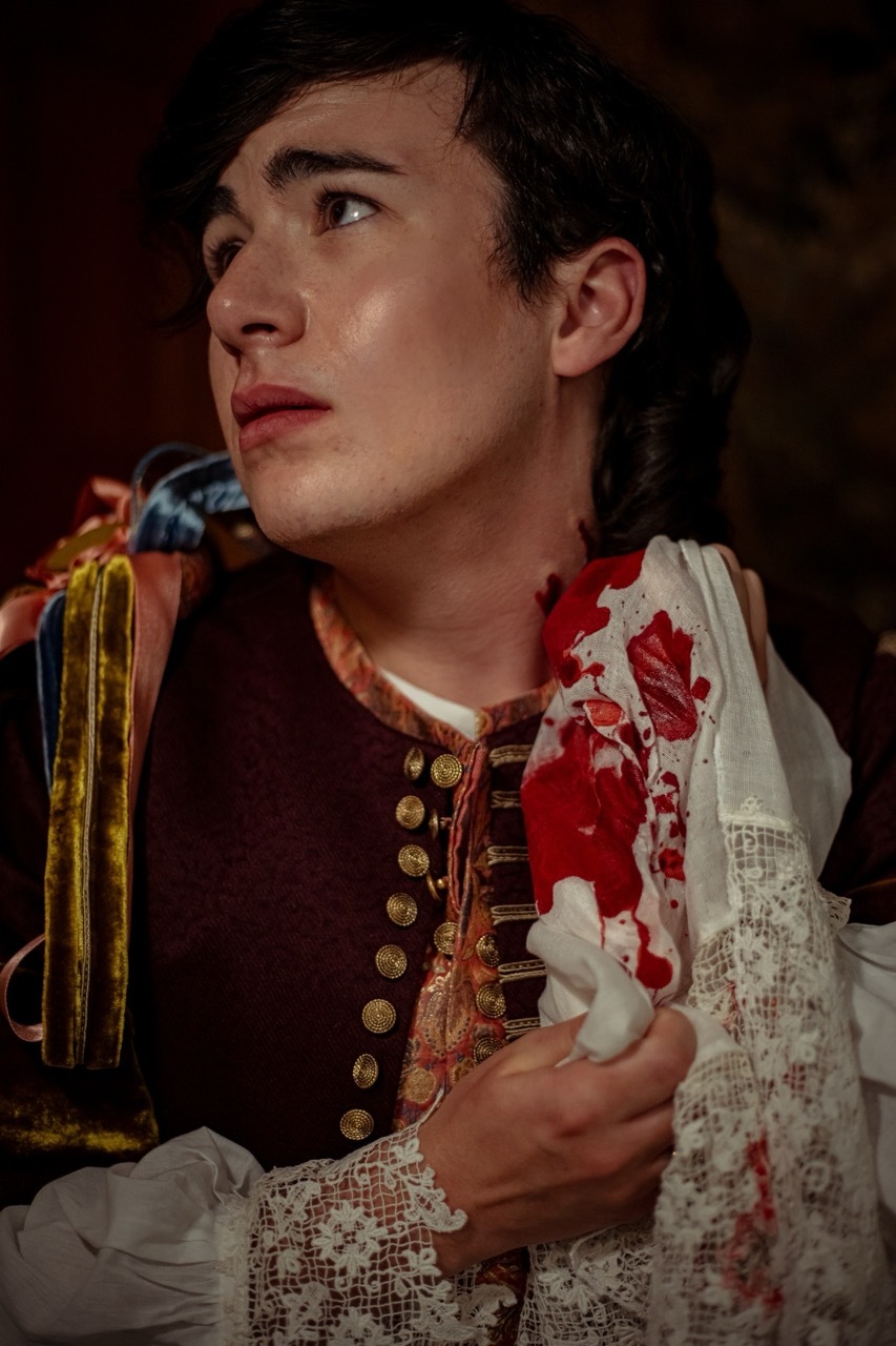 A stately man dressed in 17th-century clothing glances over his shoulder, looking dismayed. He holds a bloodied lace cravat to his neck, barely covering the vampire bite marks there.