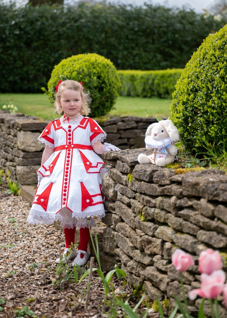 A little girl dressed in 19th-century waits by a stone wall with her little white rabbit plush. She is wearing a crisp red-and-white Victorian dress, decorated with white hearts, heart-shaped lace, and a big red bow at the back.