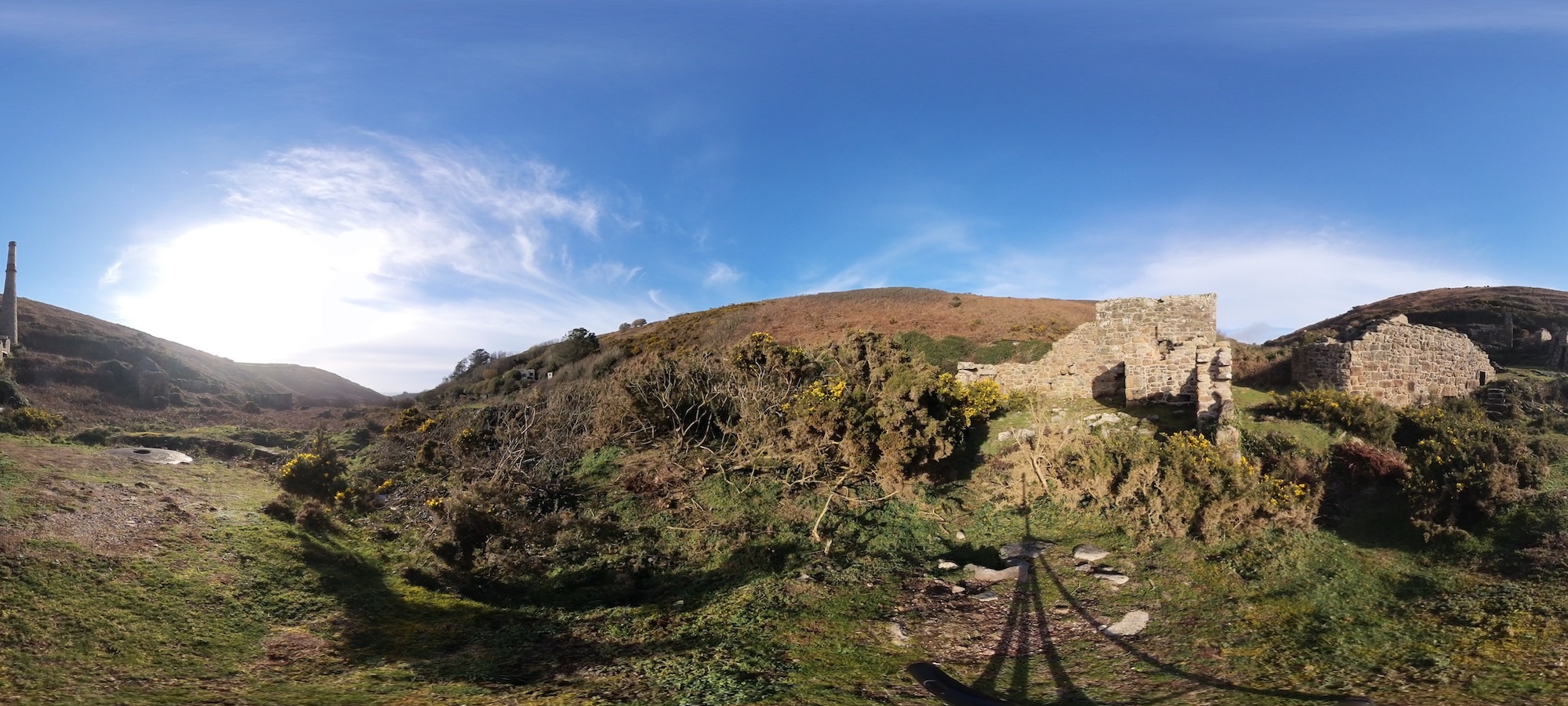 Panoramic landscape of green hills and heathland, with a cloudy blue sky above. Some stone ruins appear on the right-hand side.