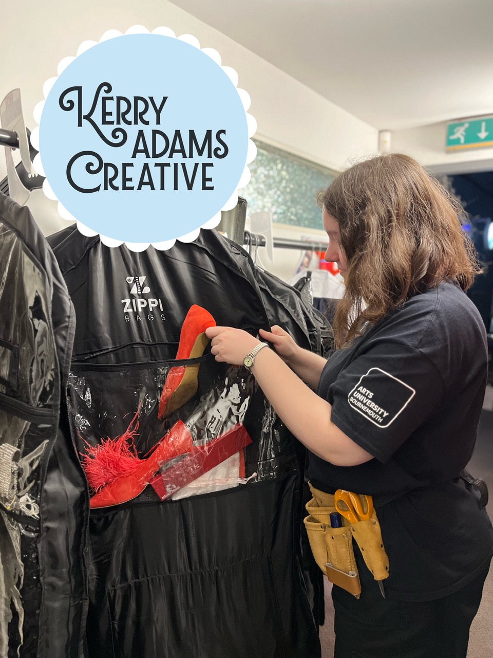 A woman with long brown hair, wearing a black t-shirt and tool belt, is arranging items with red and black fabric in a garment bag. The background shows a light-coloured wall and a sign with the text "Kerry Adams Creative".