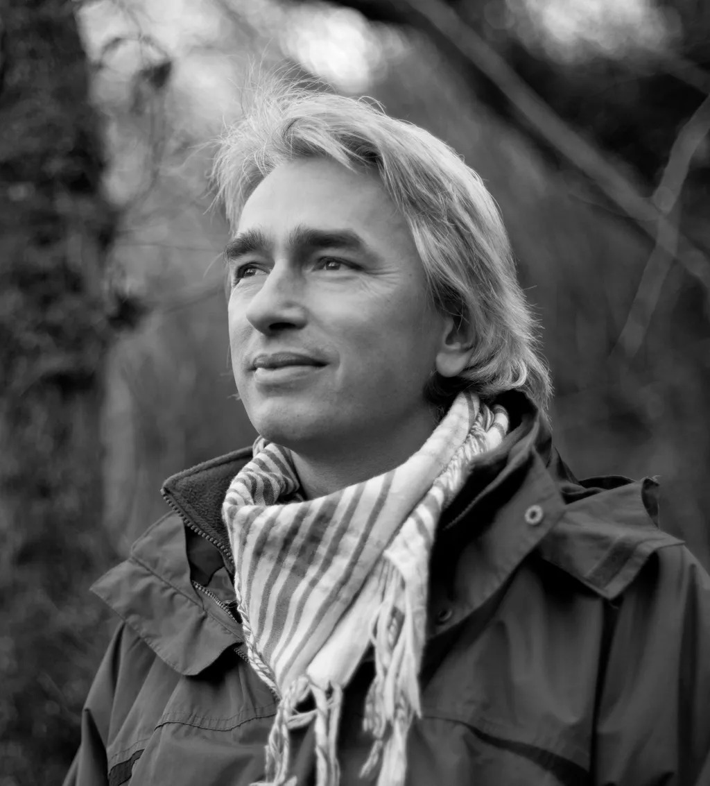 Black and white portrait of smiling woman with shoulder-length blonde hair wearing striped scarf and dark jacket, looking upward.