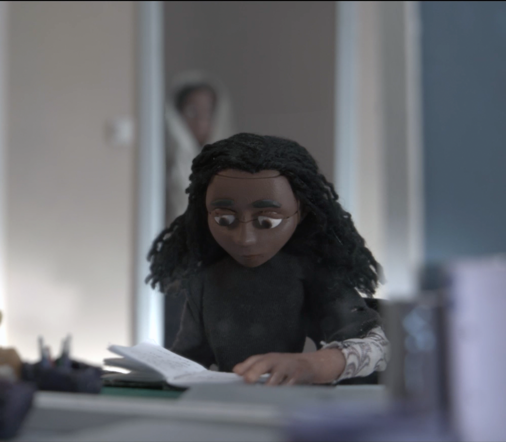 Stop motion animated character sitting at a table and reading a book with a sad expression. A blurred figure appears in the doorway behind them.