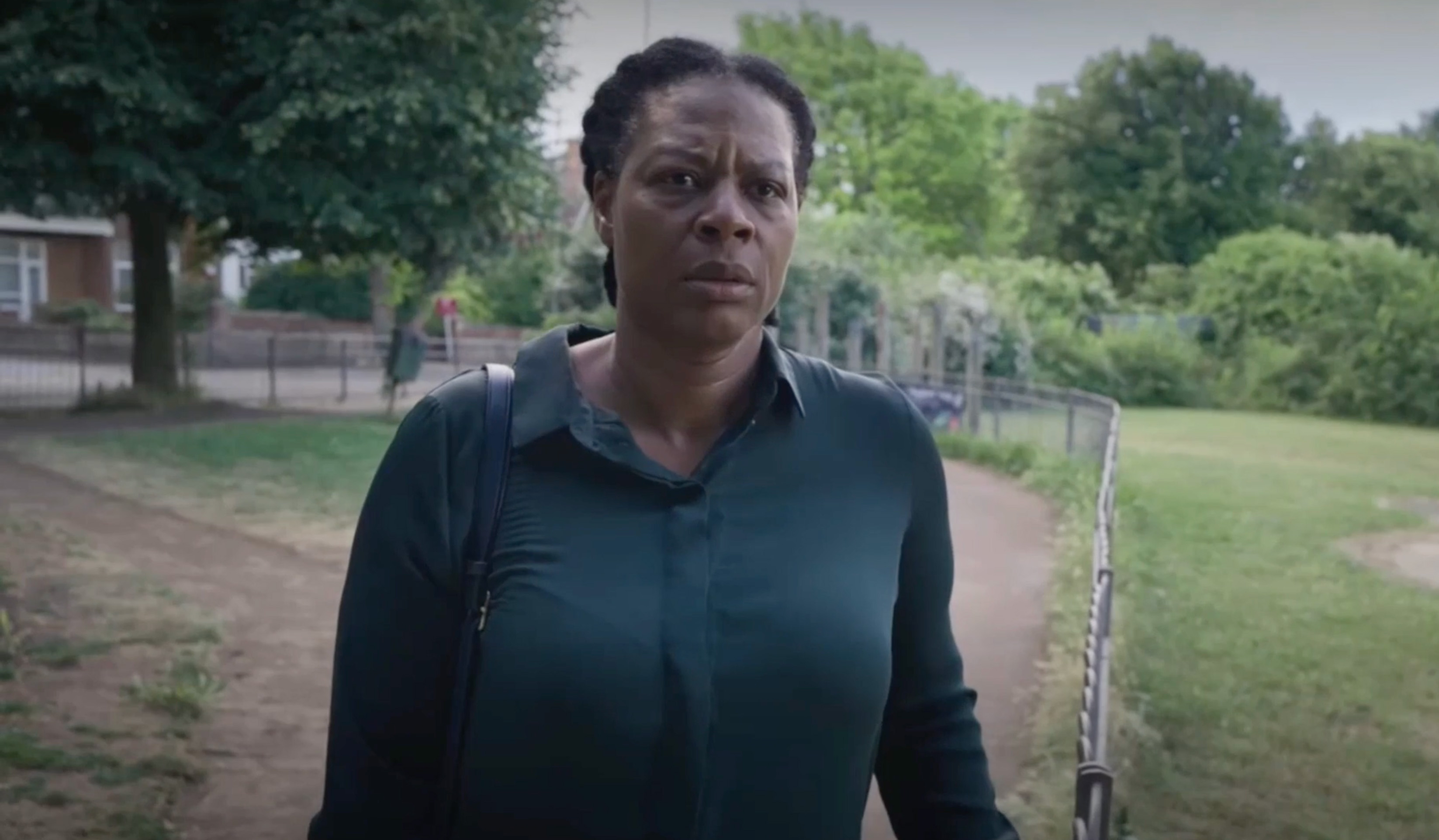 A woman in a dark green top walks along a park path, with trees, grass and a low fence in the background.