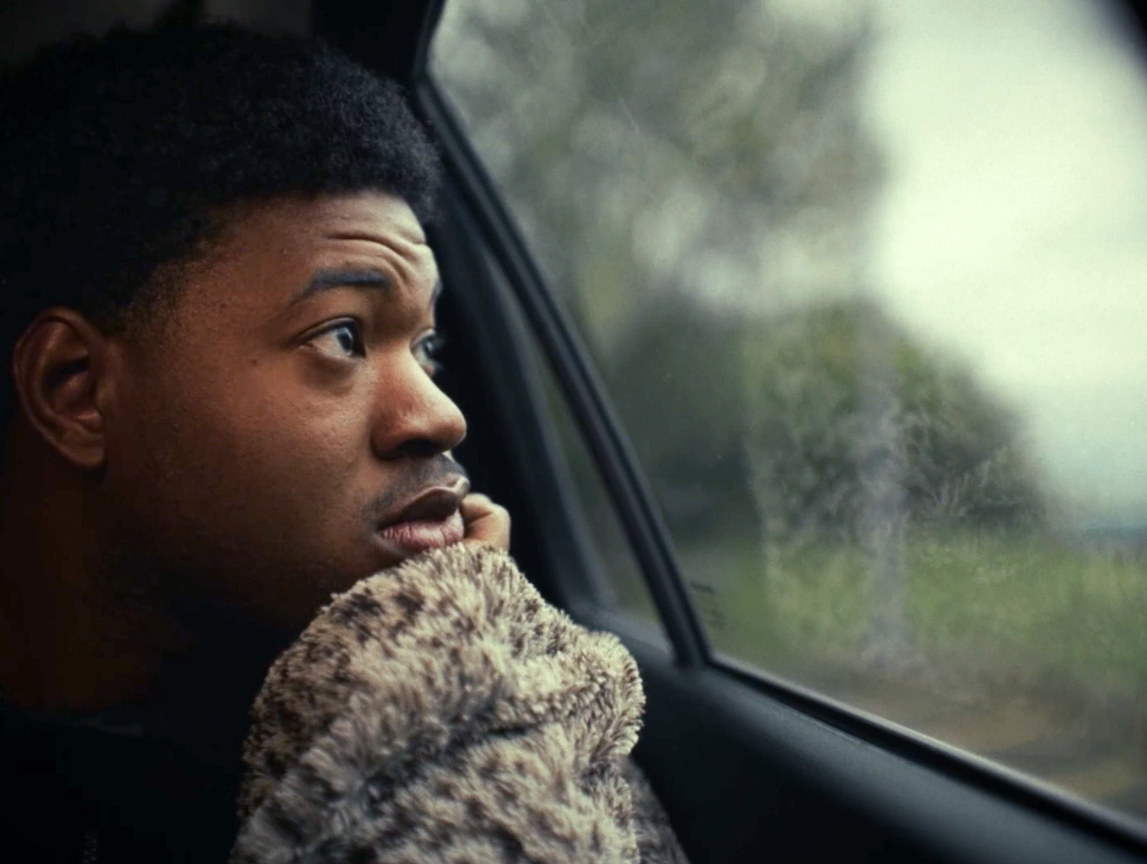 Man in chunky knit jumper sitting by car window, looking pensively to the side with blurred greenery visible outside.