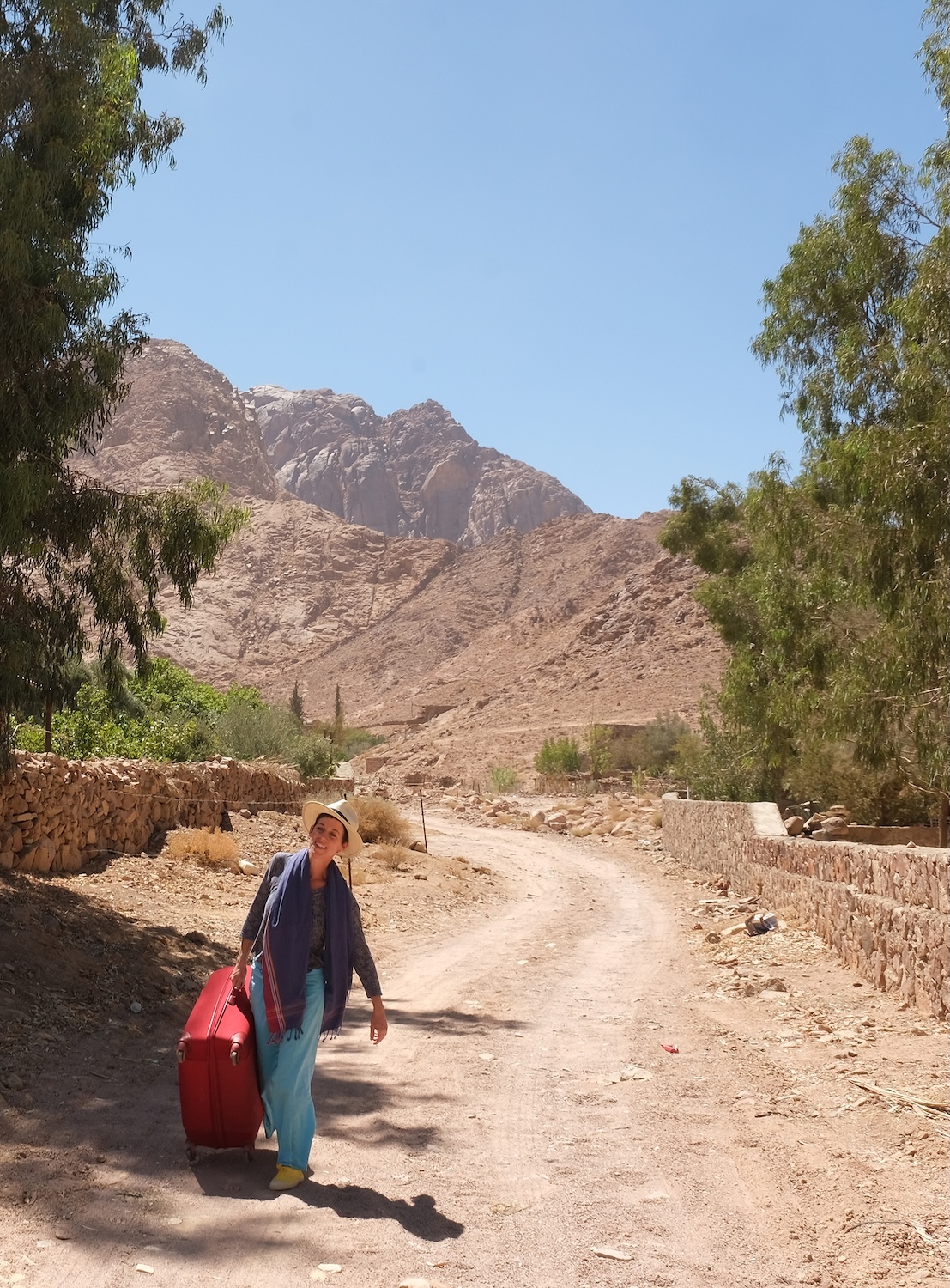 Woman in dark top and light trousers carries red bag on dusty road through desert landscape with rocky mountains and green trees.