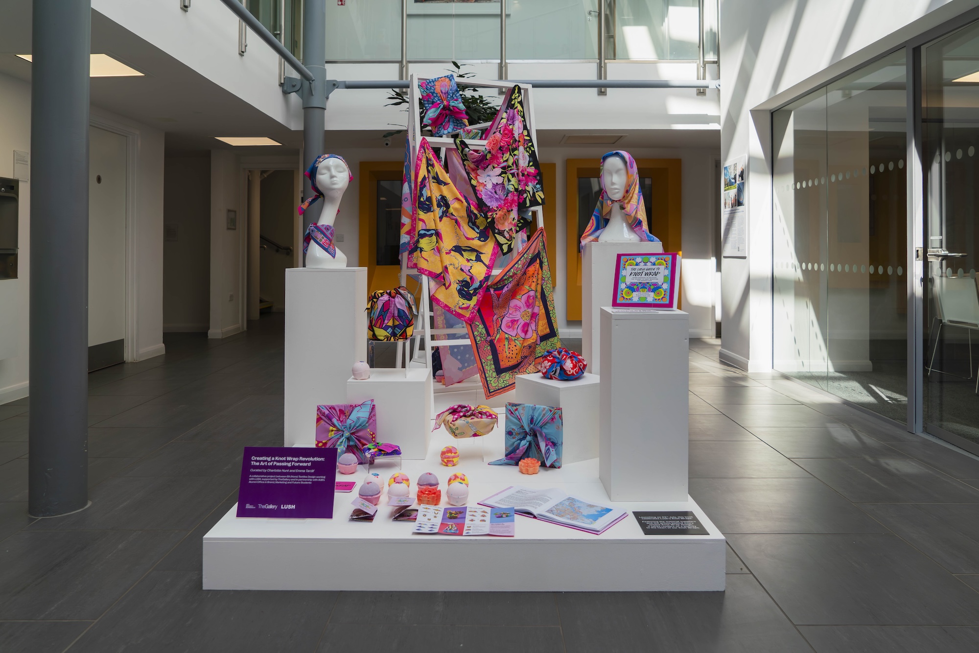 Lush Knot Wrap exhibition in South House, various multicoloured bath bombs, knot wraps and floral, multicoloured textiles on white podium and model heads.