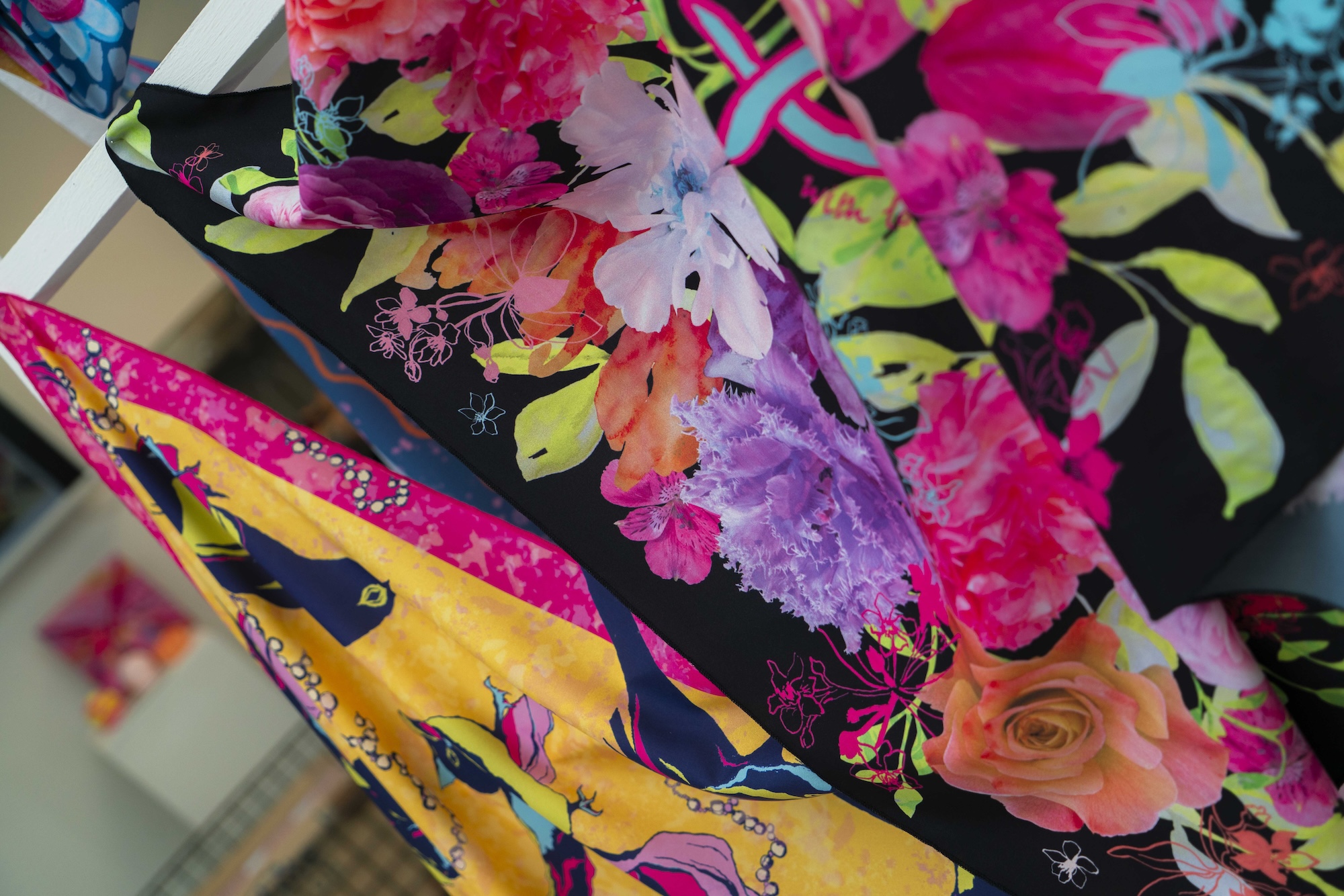 Close-up of layered, abstract, multicoloured floral textiles with plant leaves and flowers in black, green, bright pink and yellow, blurred background.