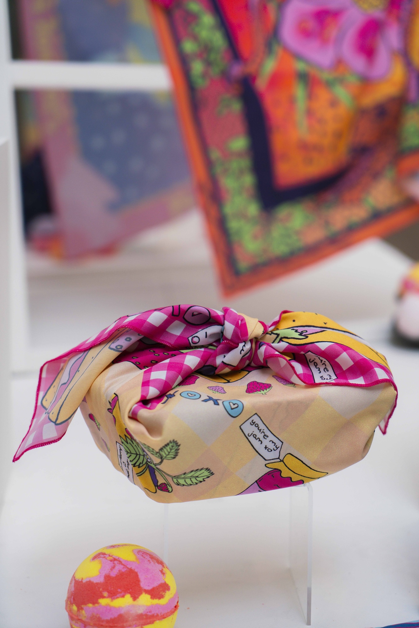 Close-up of a pink checkered and beige knot wrap with illustrations of jam and leaves, text reads 'you're my jam xoxo', blurred colourful textiles in the background.