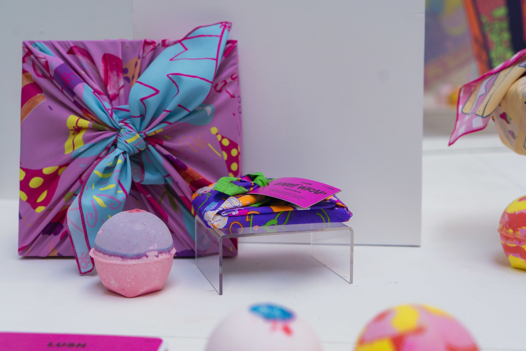 Pink, red and blue knot wrap with different patterns next to a pink-purple Lush soap and purple knot wrap, with a pink card on top. White background and blurred, colourful objects.
