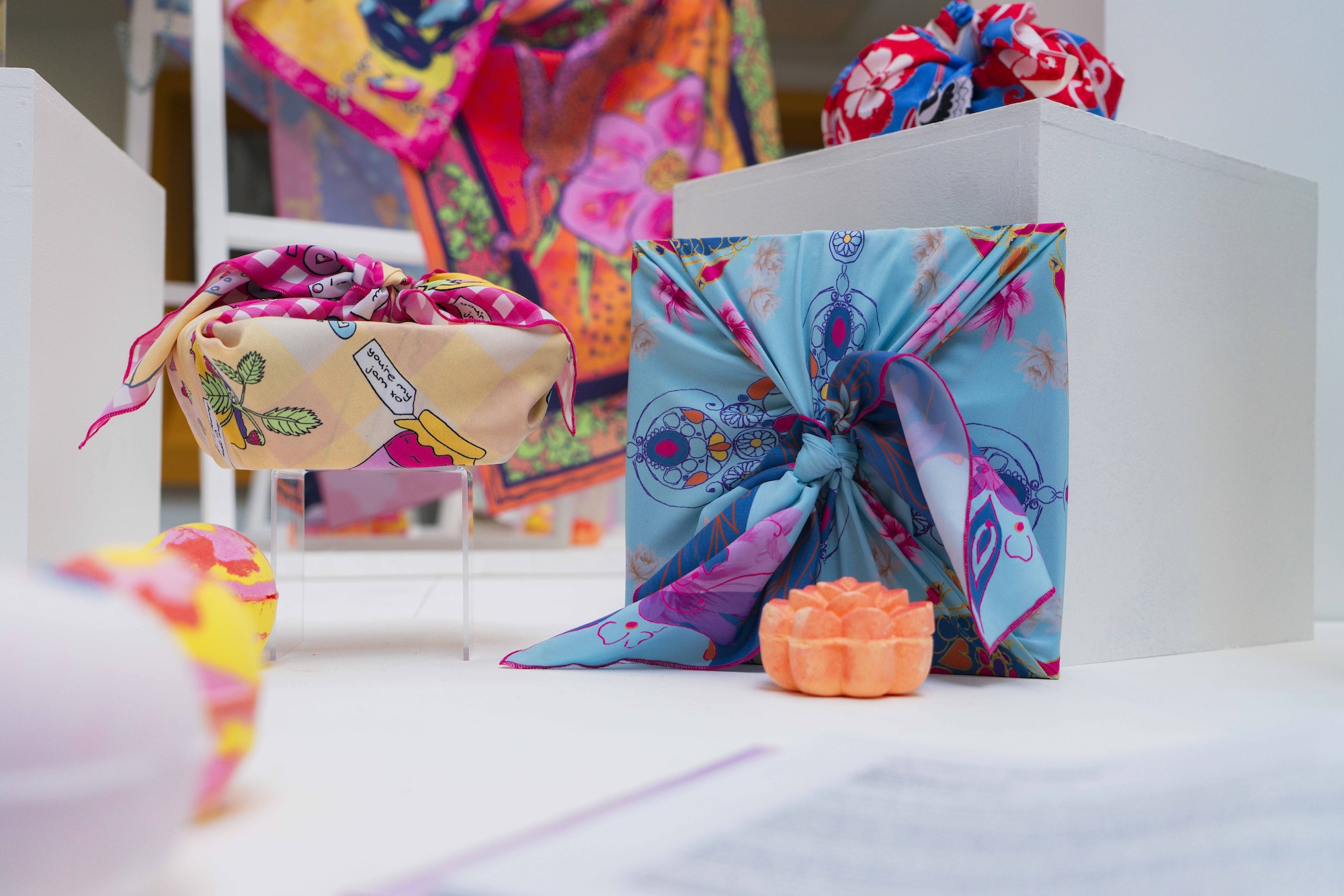 Colourful knot wraps in different shapes and patterns: one square in blue and pink behind an orange Lush soap, the other in beige with jam illustrations, blurred pink textiles in background.