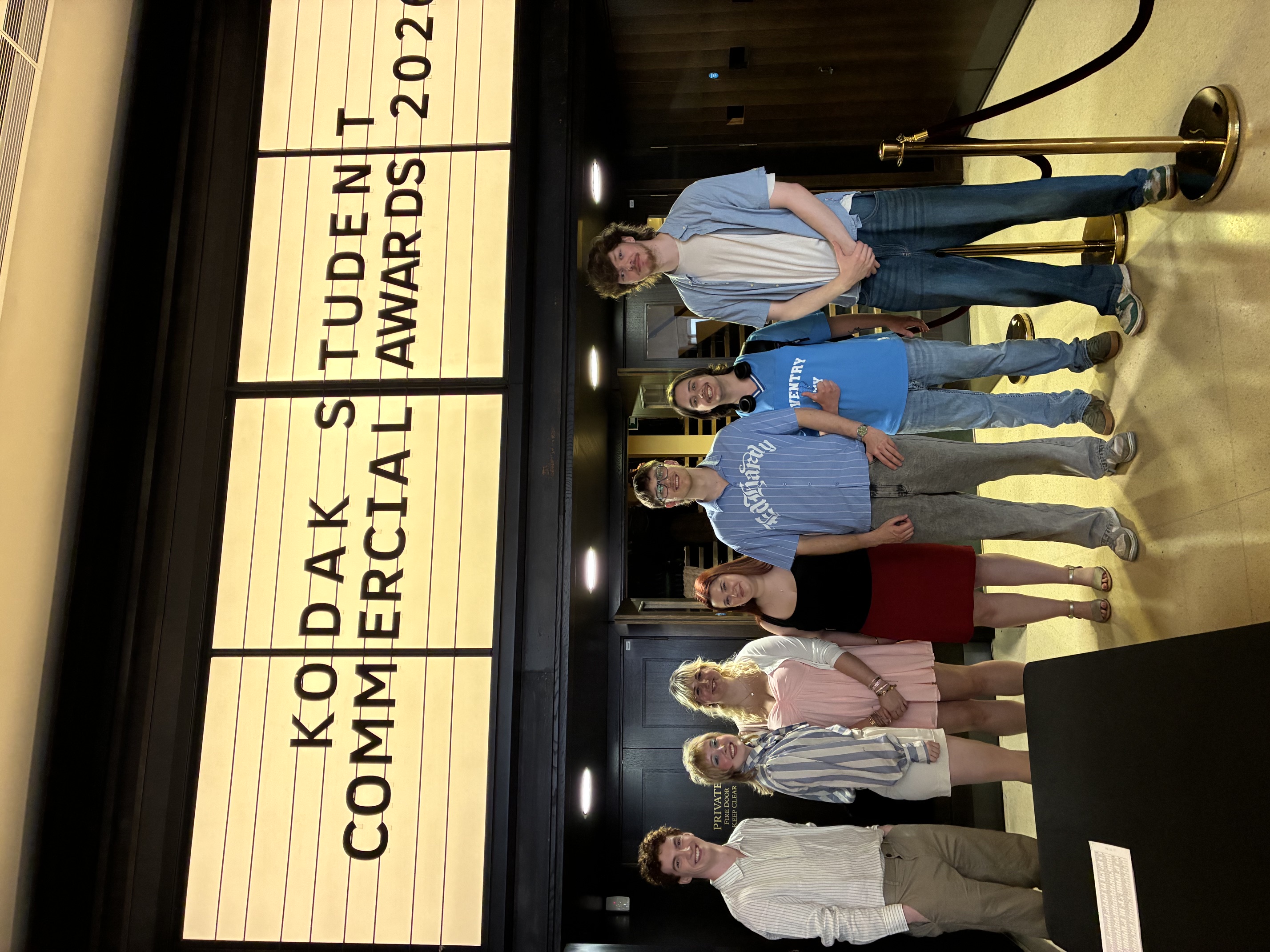 Students stood in front of yellow cinema banner with text that reads "Kodak Student Commercial Awards 2026", green carpet and doors behind them.