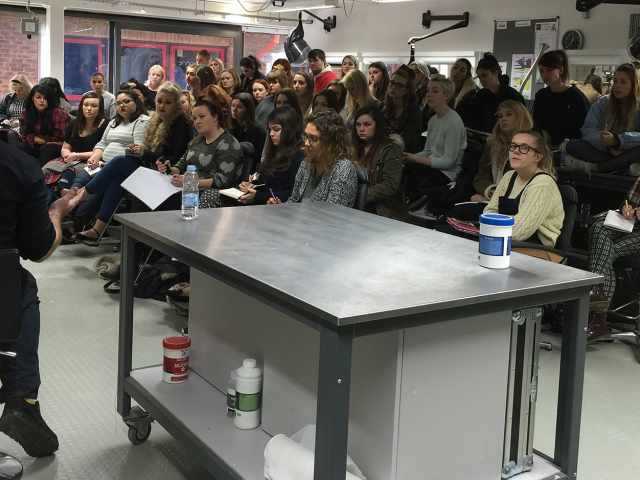 Large audience of people sitting in a studio. A large metal table is in the foreground.