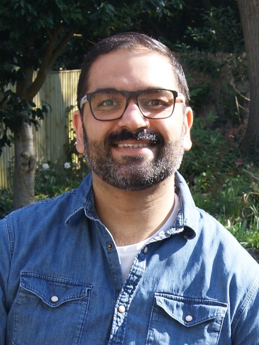 Man with dark beard and glasses wearing blue denim shirt, smiling outdoors with green foliage and wooden fence in background.