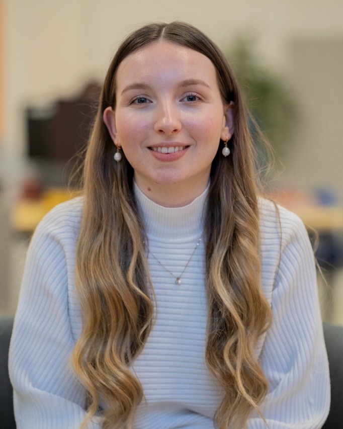 Kyra Gibson headshot with blurred background, she has long brunette hair, earrings and a light blue jumper.