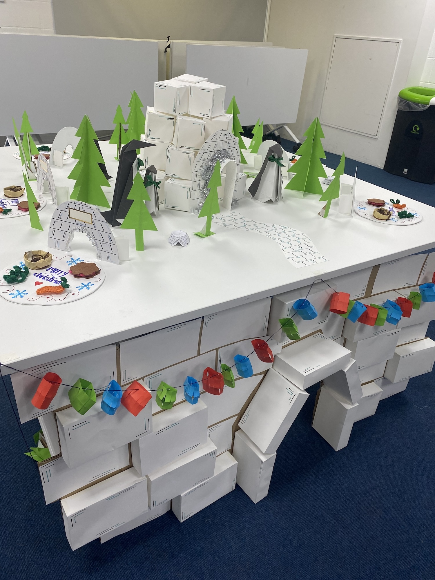 Model fireplace made from small card boxes, with a large white panel on top. A winter scene of card trees, igloos and penguins is displayed on the panel. Paper Christmas lights hang from the fireplace.