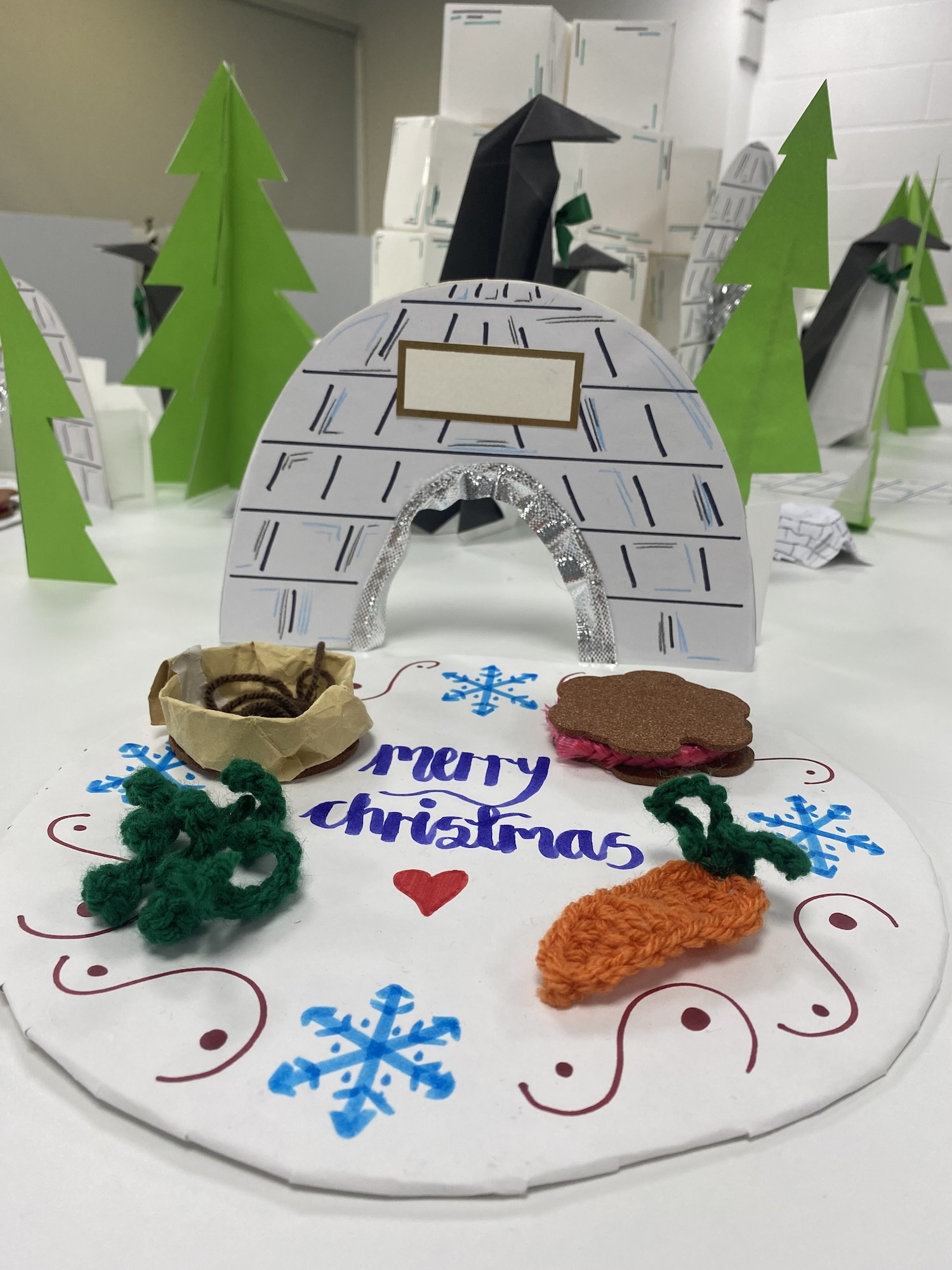 Close-up of a round paper welcome mat in front of the entrance to a card igloo. Handwritten text and decorative patterns and snowflakes appear on the mat.
