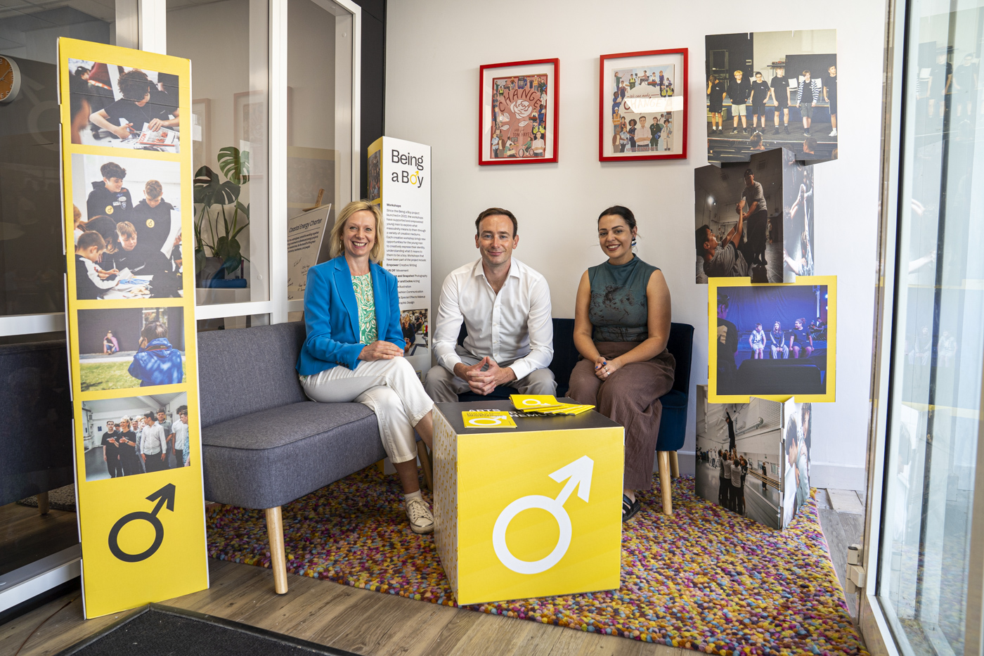 Three people are seated on a grey sofa in an office space. The woman on the left wears a blue blazer and smiles, the man in the middle is dressed in a white shirt, and the woman on the right is in a green top. Yellow banners with images and text about 'Being a' are displayed nearby.
