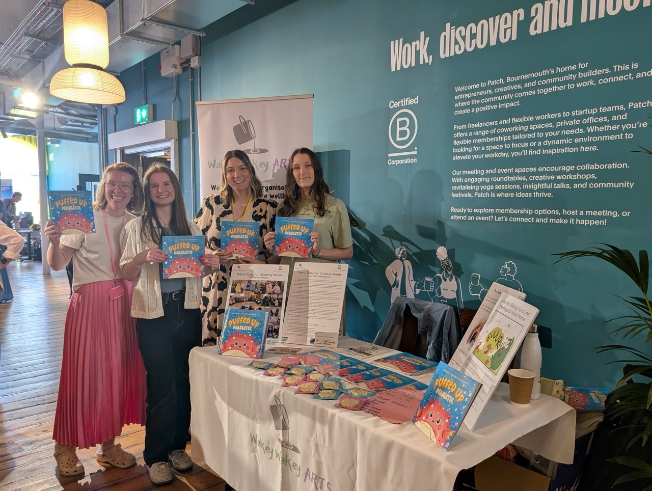 From left to right: Claire Reilly, Emma Watremetz, Becky White and Elana Evans stood behind a booth with copies of 'Puffed Up Pearlette', teal wallpaper background.