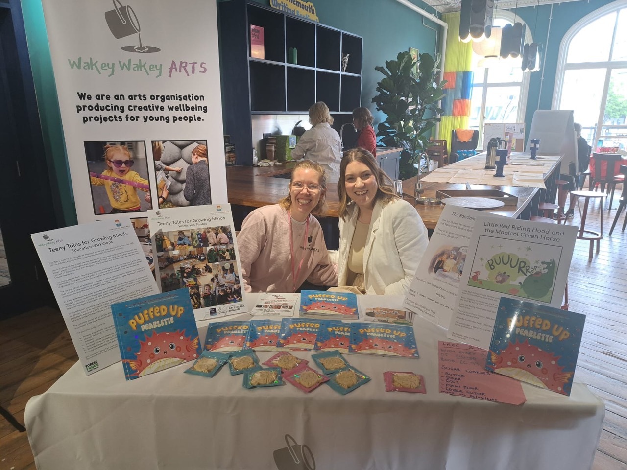 Claire Reilly and Becky White sat by a stand and banner with copies of 'Puffed Up Pearlette' spread out.
