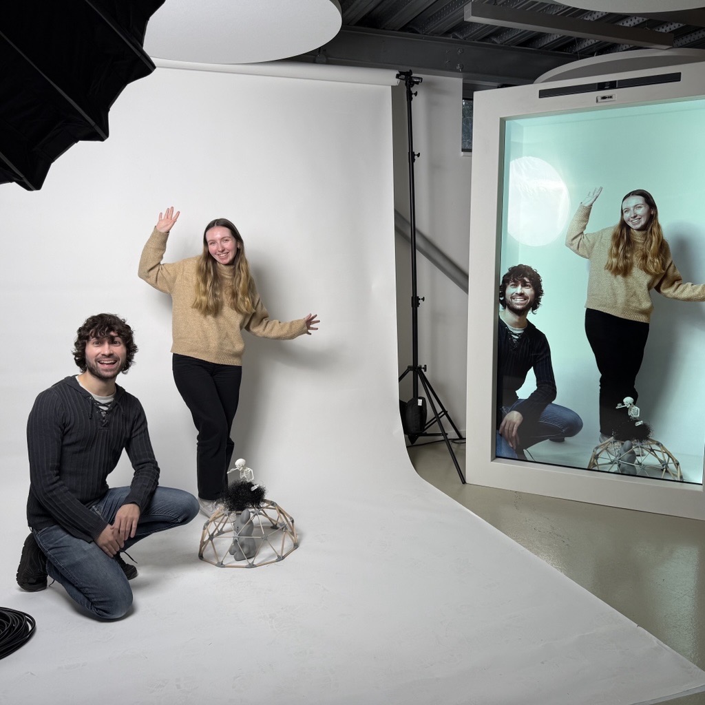 Oliver Moore Oats and Kyra Gibson stood in front of a white background to the left, to the right, a projection of them in a holographic box.