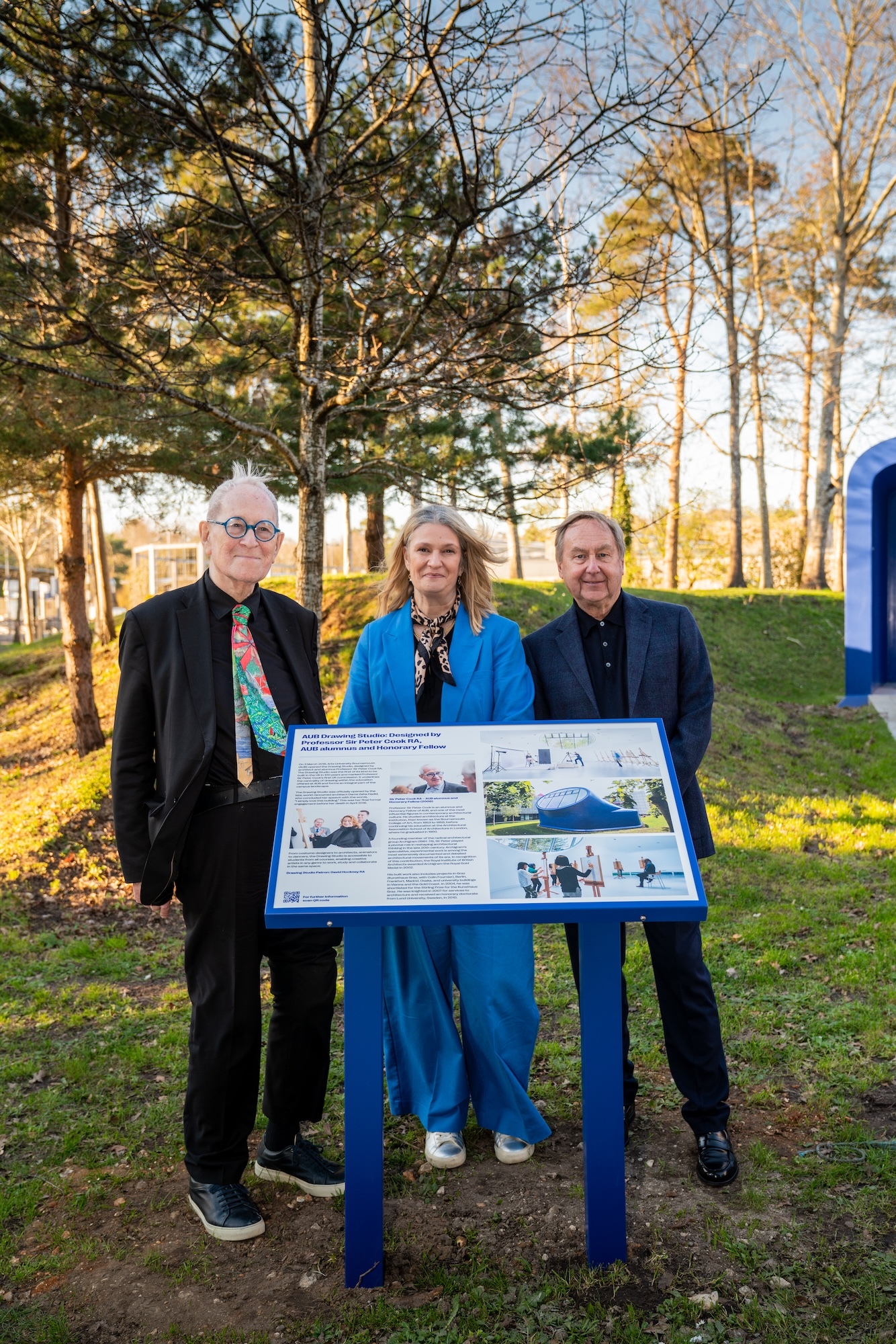 Three people standing behind blue information board in park setting with autumn trees and grass in background.