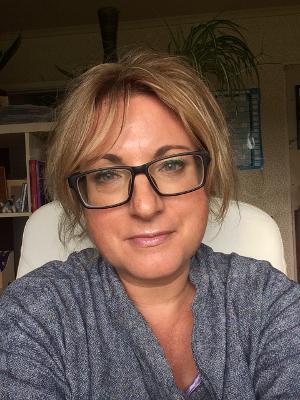 Woman with blonde hair and black-rimmed glasses wearing grey top, sitting indoors with bookshelves visible in background.