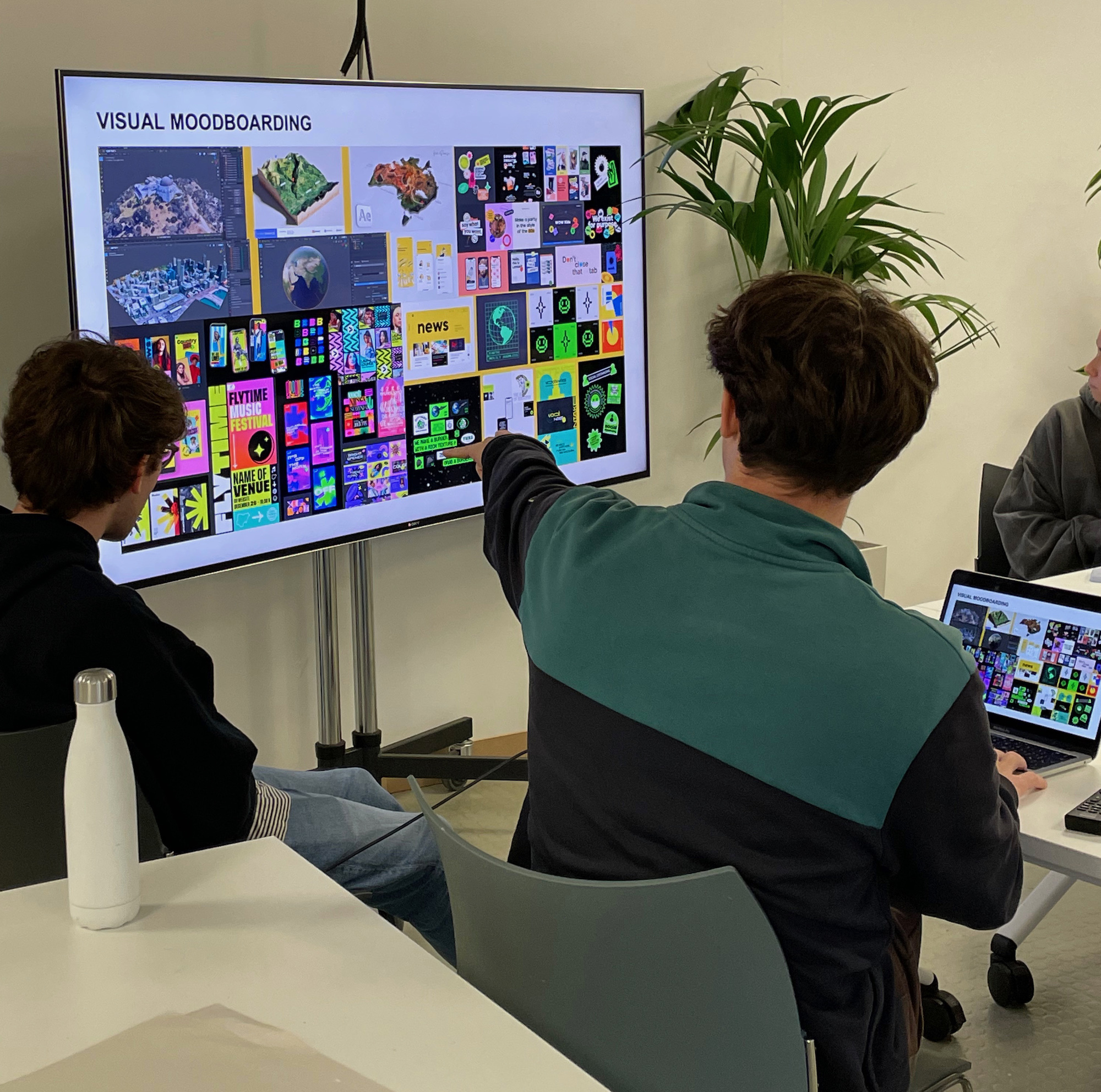 Two BA (Hons) Graphic Design students viewed from the back; one of them is pointing at a screen with a colourful mood board on it.