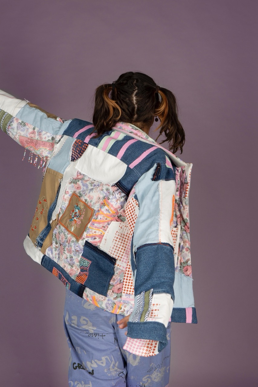 Person with dark hair in pigtails turned away from the camera, wearing patchwork jacket with blue, pink, white, and floral fabric squares against purple background.