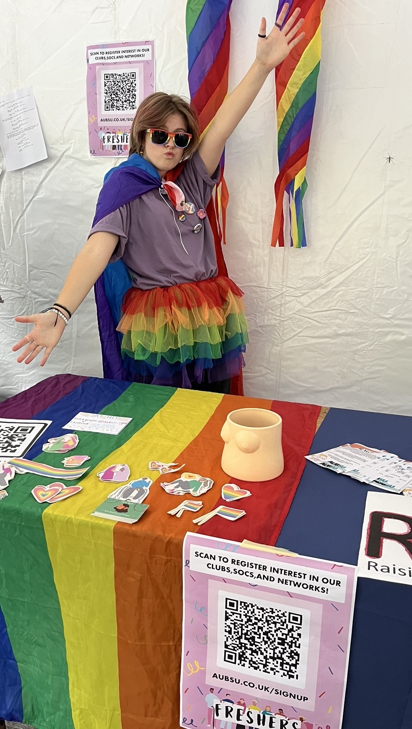 A person in costume poses in front of a table with rainbow banners and ribbons in front and behind