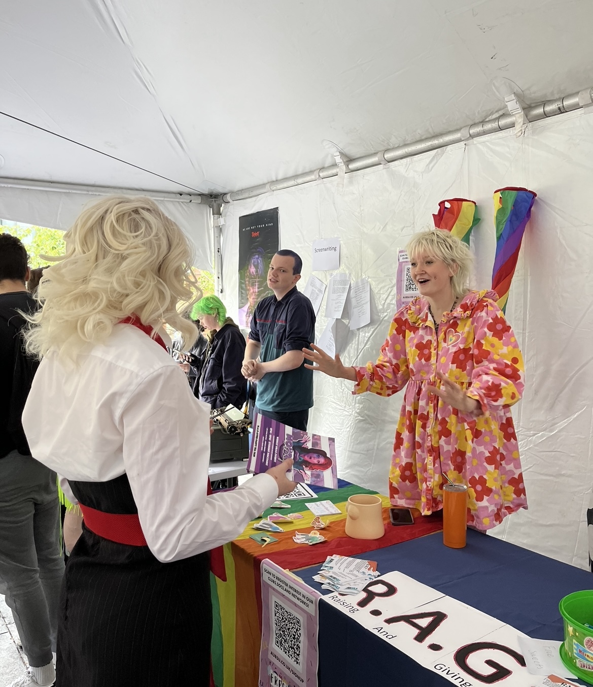 Two people chat across a table with a rainbow flag across it. Other people in the background are watching.