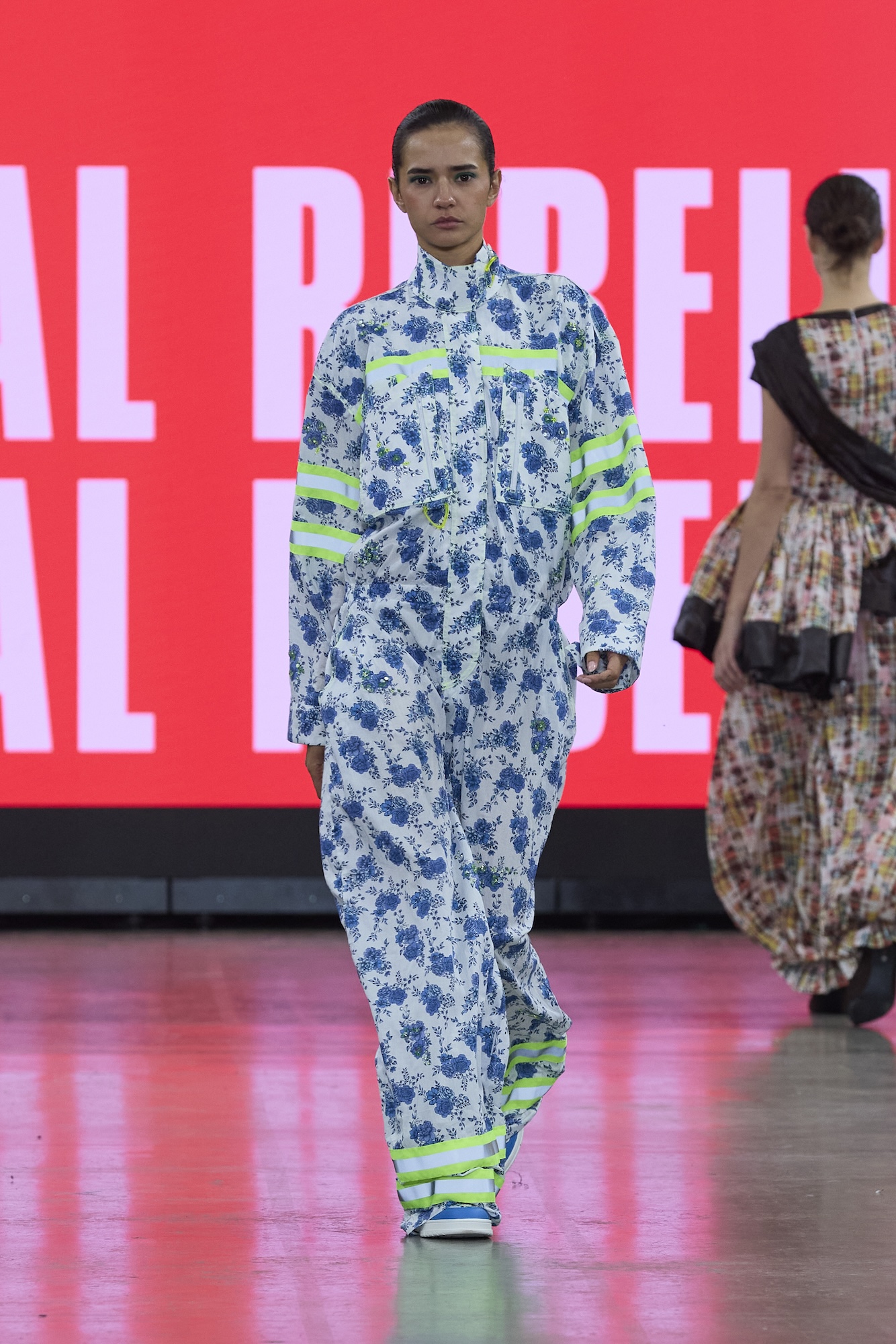 A model in a blue and white floral outfit with neon green stripes walks on a runway, with a red illuminated backdrop and another model in patterned clothing behind her.