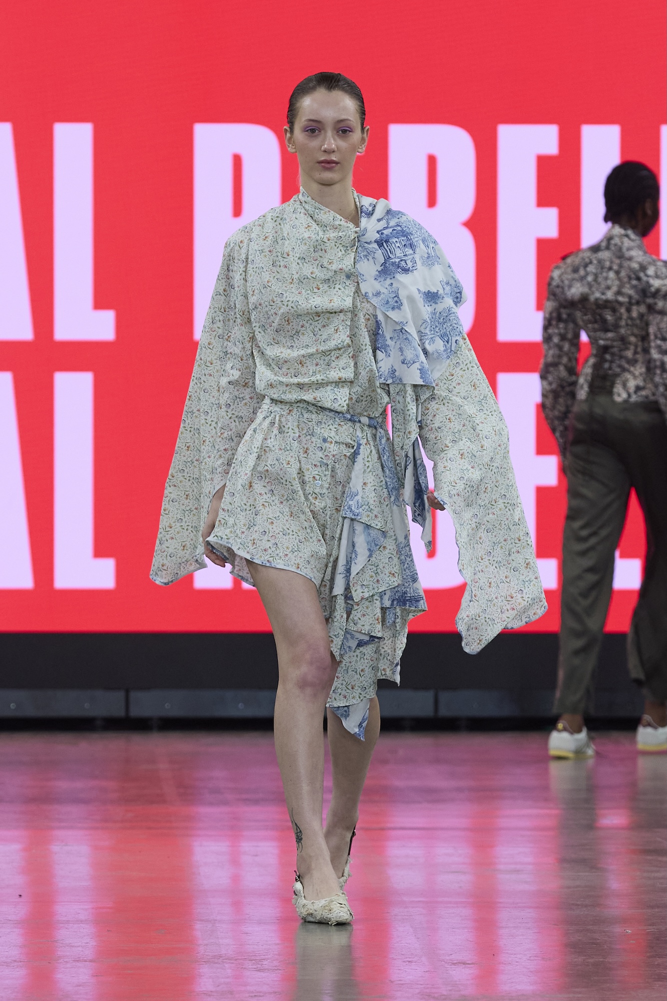 A model in a pale blue patterned asymmetrical dress walks a runway against a bright red backdrop with large white lettering.