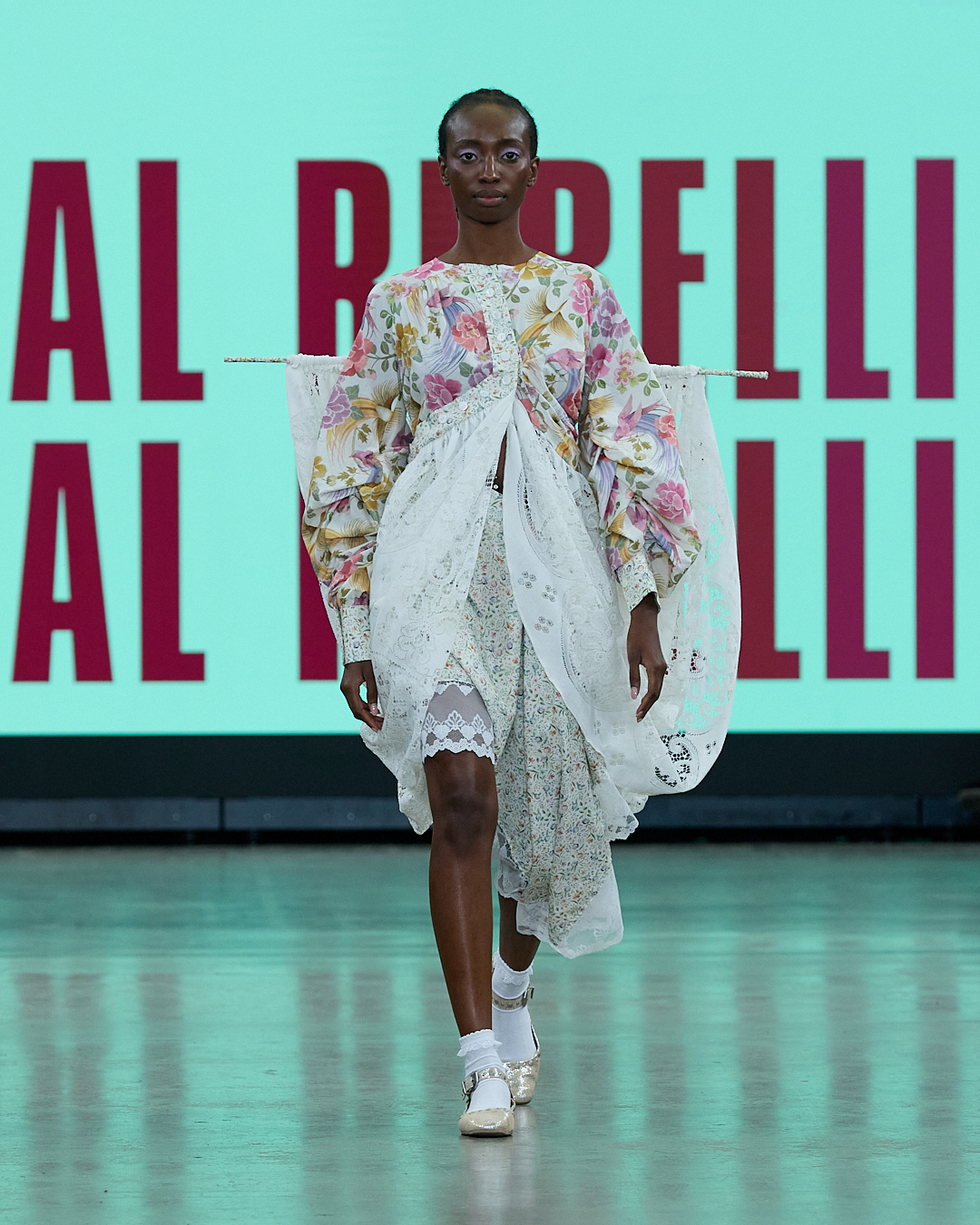 A model wears a floral and lace dress with wide sleeves on a runway, with a turquoise backdrop behind her.