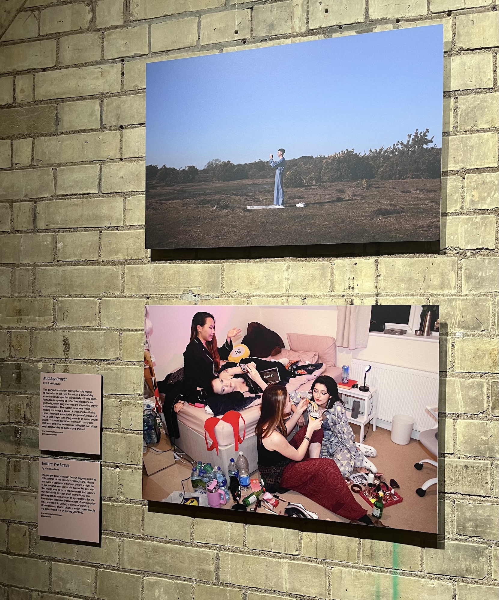 Two framed photographs hang on a brick wall, showing a person standing in a barren landscape above and several people seated on the floor of a room below.