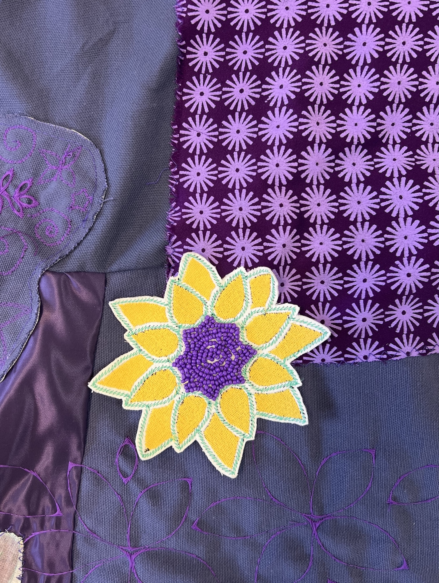 Textile artwork with purple patterned fabric, grey and purple fabrics, and yellow sunflower appliqué with purple centre and white outline.