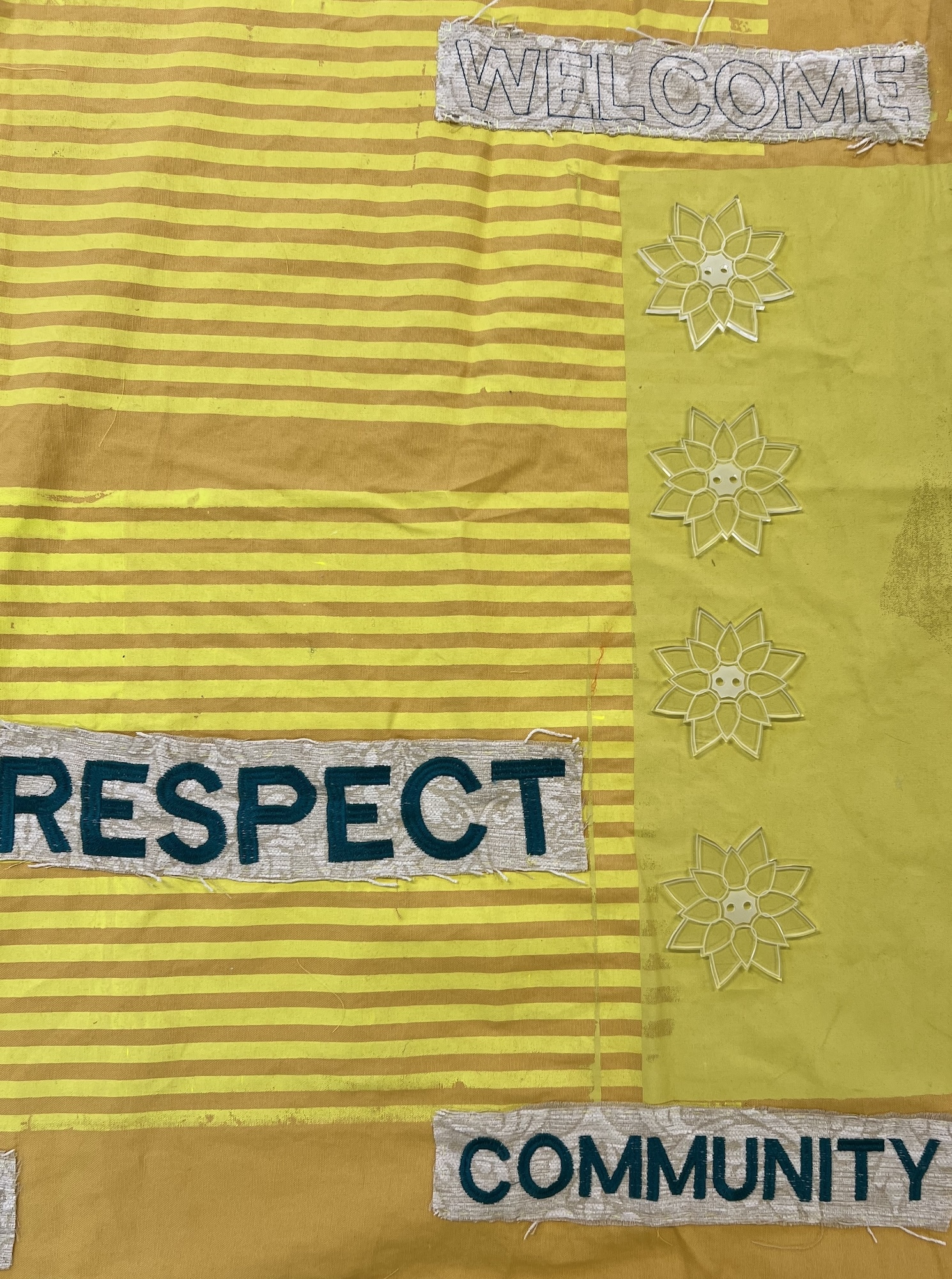 Yellow fabric with horizontal orange stripes, four white flower shapes arranged vertically, and three grey text labels reading "WELCOME", "RESPECT", and "COMMUNITY".