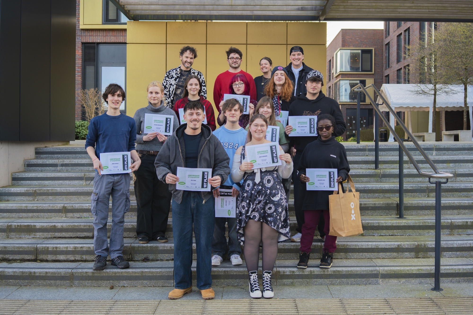 Group of BA (Hons) Graphic Design students and staff holding certificates on concrete steps outside Campus Halls at Arts University Bournemouth with metal railings.