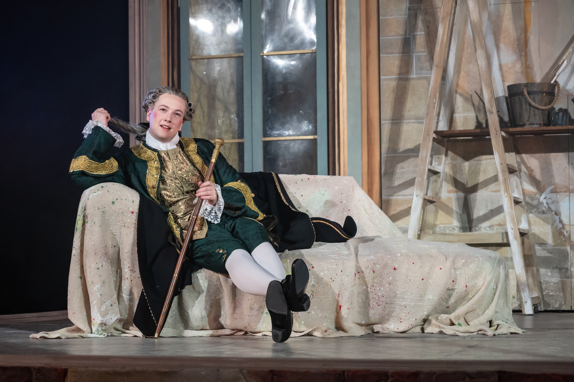 A BA (Hons) Acting student in 18th century-style costume and holding a cane sits on a sofa, performing in La Casa Nova.