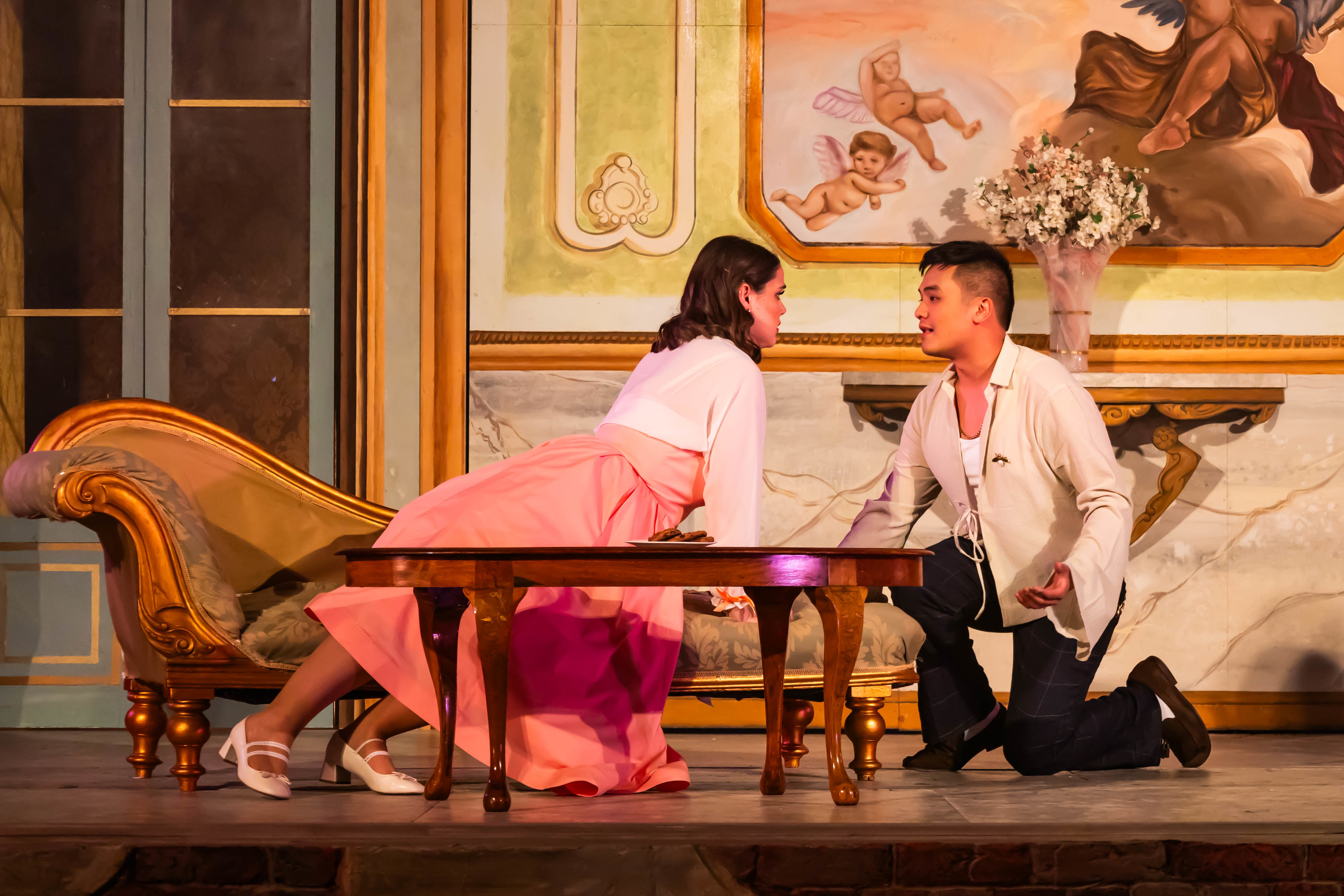 Two BA (Hons) Acting students performing a scene from La Casa Nova at Palace Court Theatre. The student on the left is leaning over a table, while the one on the right kneels down opposite her.