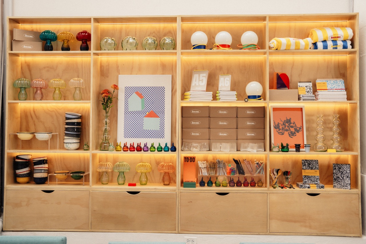 A wall of wooden shelving units displays colourful glassware, books and small decorative items, with warm встроенный lighting and drawers along the bottom.