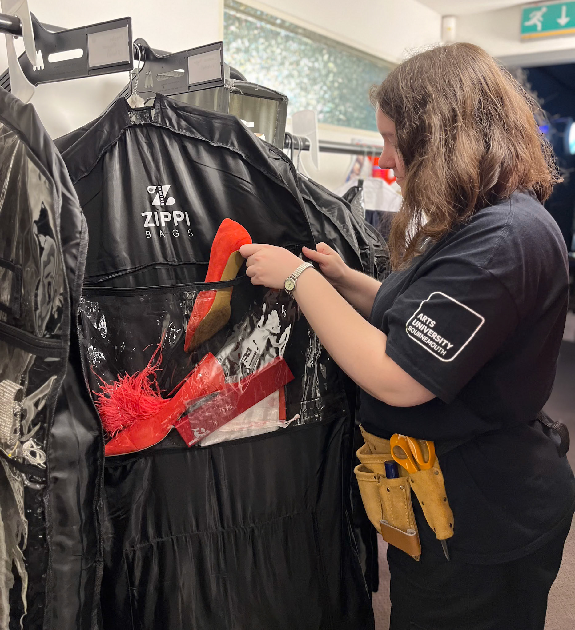 A woman in a black t-shirt examines items inside a Zippi garment bag hanging on a rack. The bag contains a red feathered item and a pair of high-heeled shoes. She wears a tool belt at her waist, indicating a costume or theatre setting.